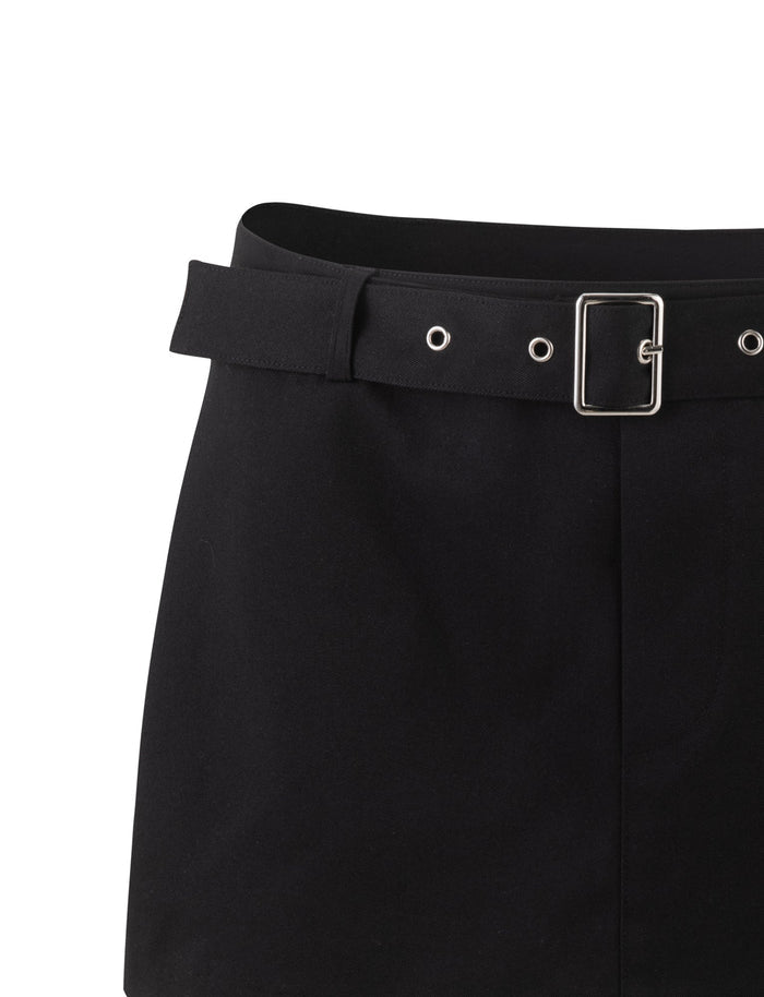 Close-up of the NADINE SKORT - BLACK by TIGERMIST, featuring a matching black belt with silver grommets and a rectangular buckle. The fabric appears to be smooth and plain, extending downward from the belted waist area.