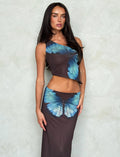 A woman with long dark hair stands against a textured gray background. She is wearing a stunning TIGERMIST two-piece outfit featuring the NANCI SKIRT - BROWN : BUTTERFLY and a sleeveless top, both decorated with large, realistic blue butterfly prints. She gazes confidently at the camera.