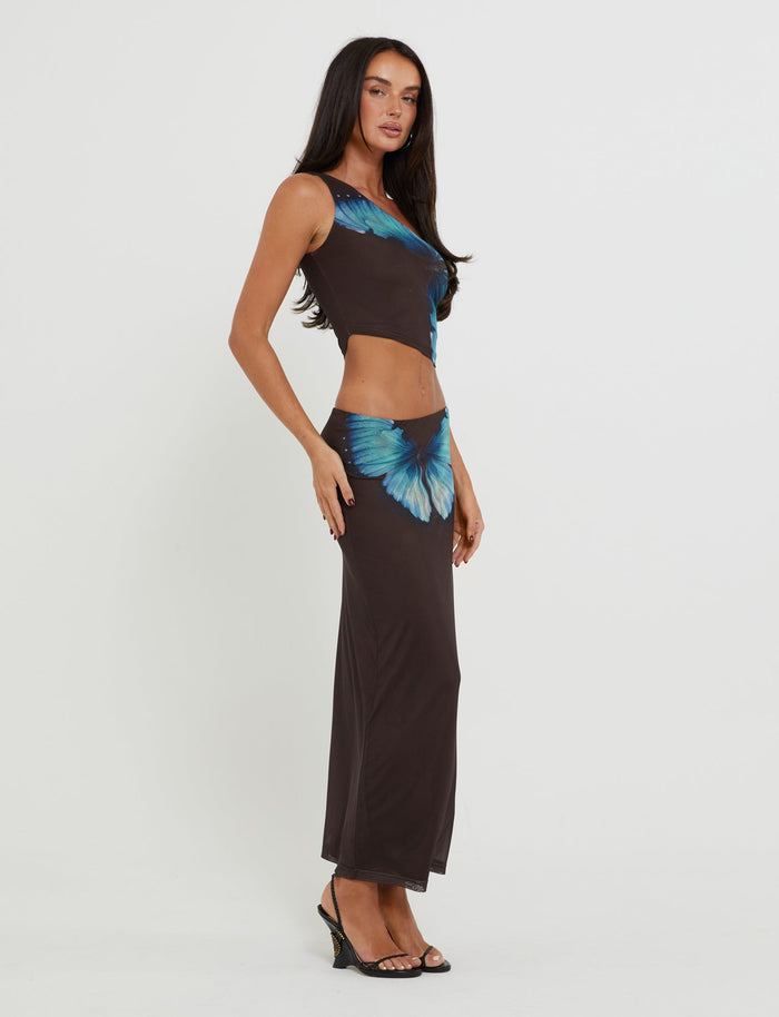 A woman with long dark hair is wearing the form-fitting NANCI Skirt - Brown : Butterfly from TIGERMIST, featuring blue butterfly patterns on the top and hip area. Posed sideways to the camera, she stands with one hand resting on her thigh and is wearing black high-heeled sandals.