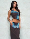A woman with long dark hair is wearing the NANCI TOP - BROWN : BUTTERFLY from Tigermist, paired with a matching long skirt. The outfit features an asymmetrical design and dark fabric adorned with a blue butterfly pattern. She stands against a textured, light-colored wall, posing with one hand on her hip and the other at her side.