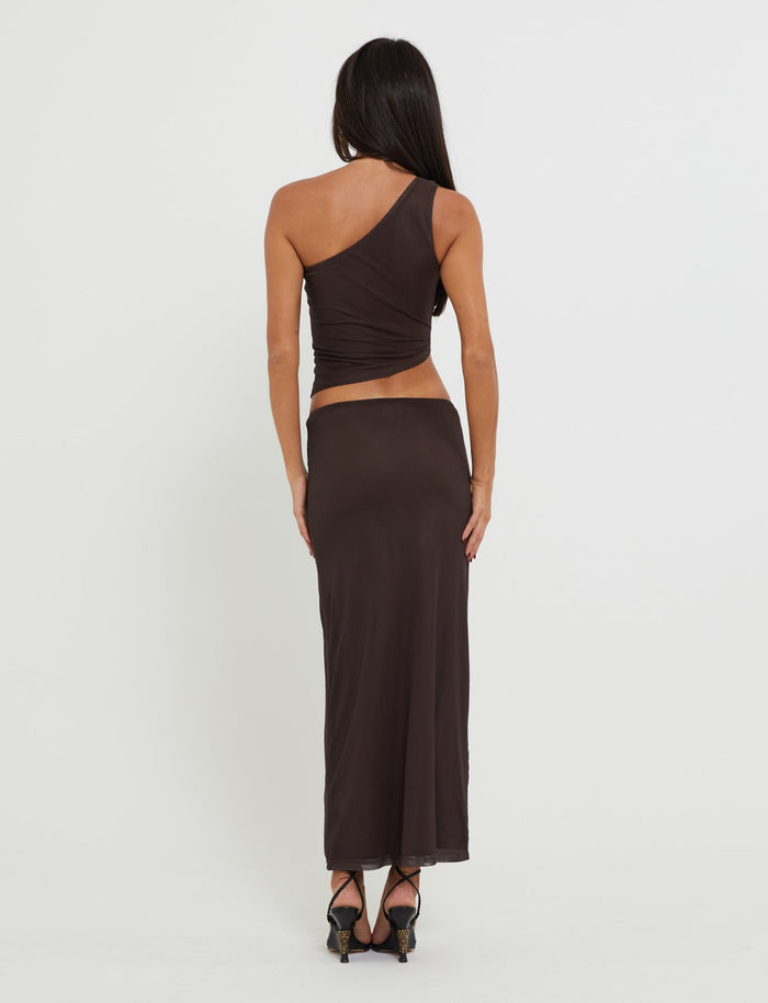 A woman with long dark hair stands with her back to the camera, wearing a chic one-shoulder brown top named NANCI TOP - BROWN : BUTTERFLY from TIGERMIST and a matching black maxi skirt. She completes her look with black high heels, set against a plain white background.