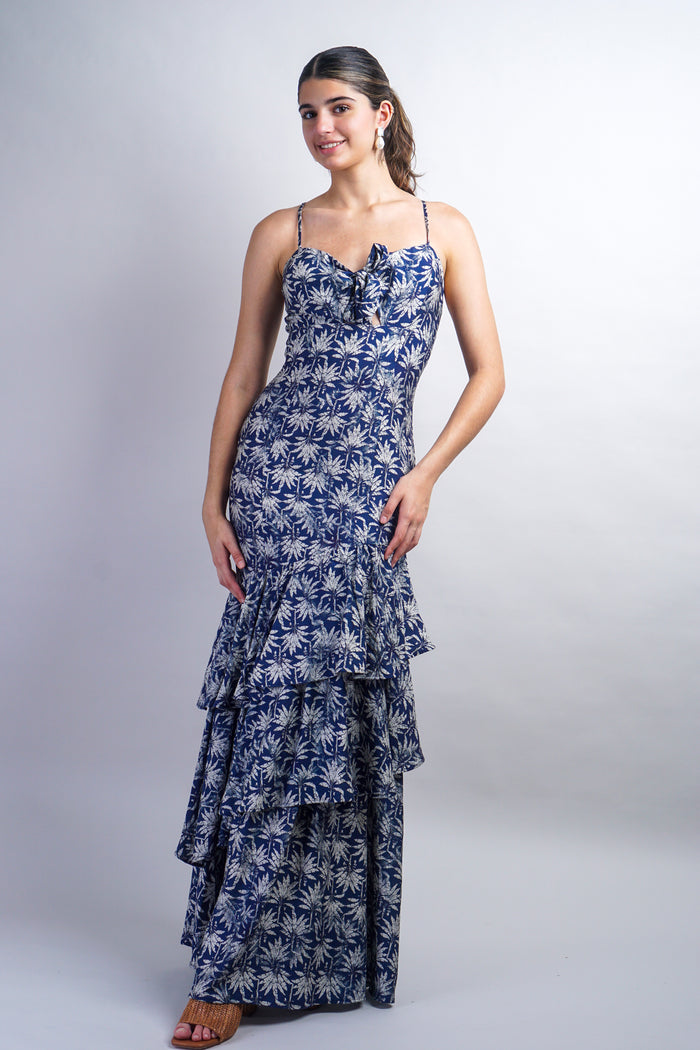 A woman modeling a sleeveless, tiered maxi dress with a blue and white palm tree print against a light gray studio background.