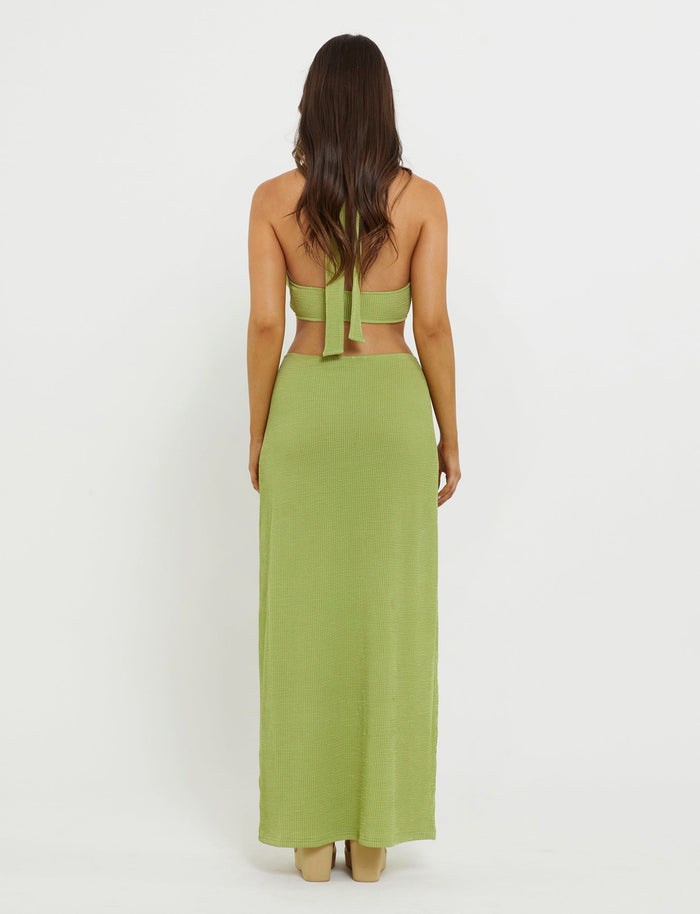 A person with long dark hair is facing away from the camera, wearing a TIGERMIST light green halter-neck top and the NEERA MAXI SKIRT in lime green, paired with beige high-heeled shoes. The background is a plain white wall.