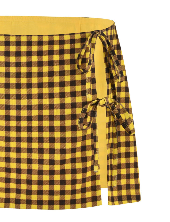 A close-up image of the NICOLA SKIRT - YELLOW : CHECK from TIGERMIST, featuring a yellow and brown checkered pattern with two side ties that reveal a slightly open side, showcasing the inner yellow fabric.