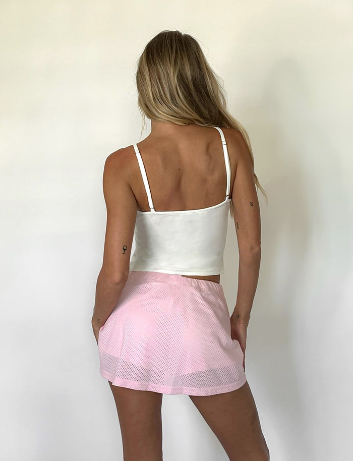 A person with long blond hair stands facing away from the camera, wearing a stylish white tank top and the NYDIA SKORT in Blush Dusty Pink from TIGERMIST. They have tattoos on their arms and are posing against a plain white background.