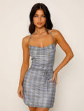 A woman stands against a plain background, wearing a TIGERMIST PATRA DRESS - MULTI : MIRO CHECK. The sleeveless dress features a plaid pattern in shades of blue and gray, with thin straps and a fitted waist that includes slight gathering. She has long, wavy dark hair and maintains a neutral expression.