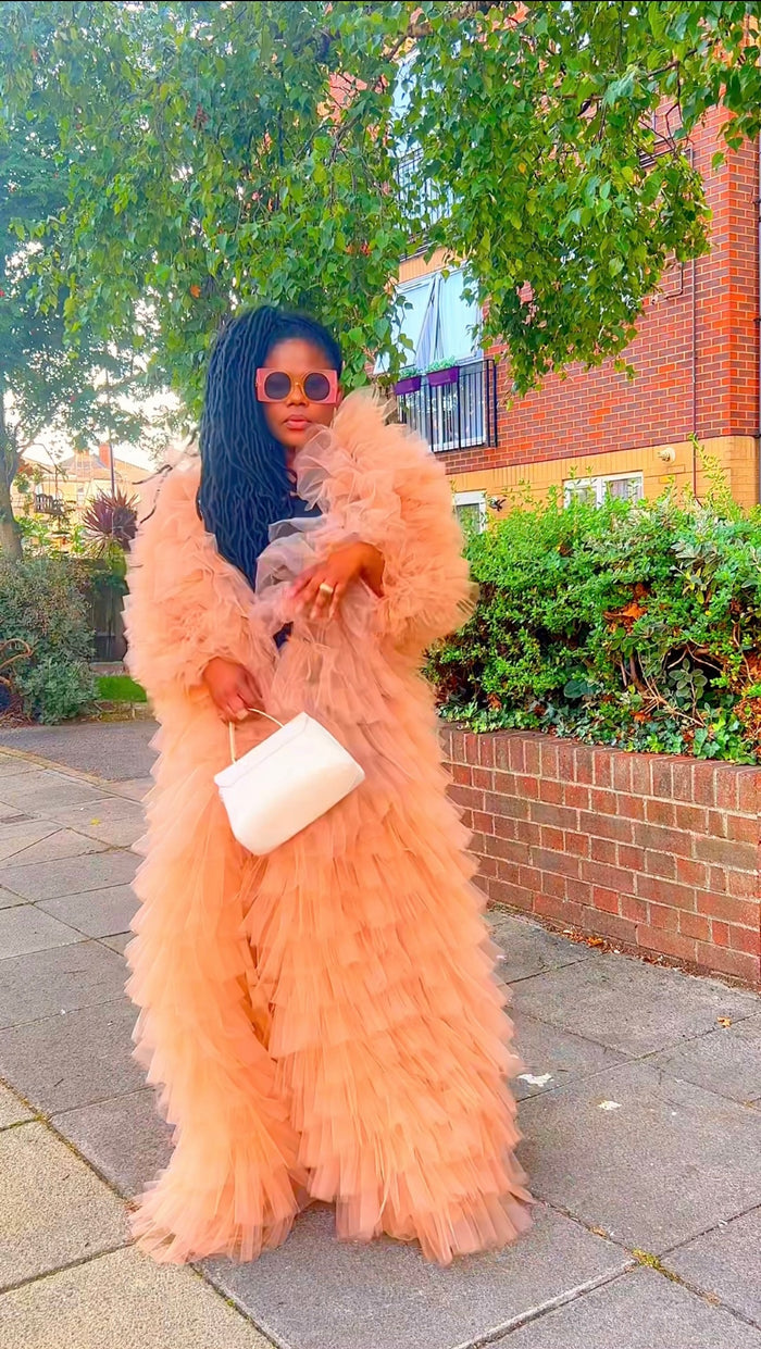 Woman in a peach fur coat standing on a sidewalk with greenery and a building in the background.