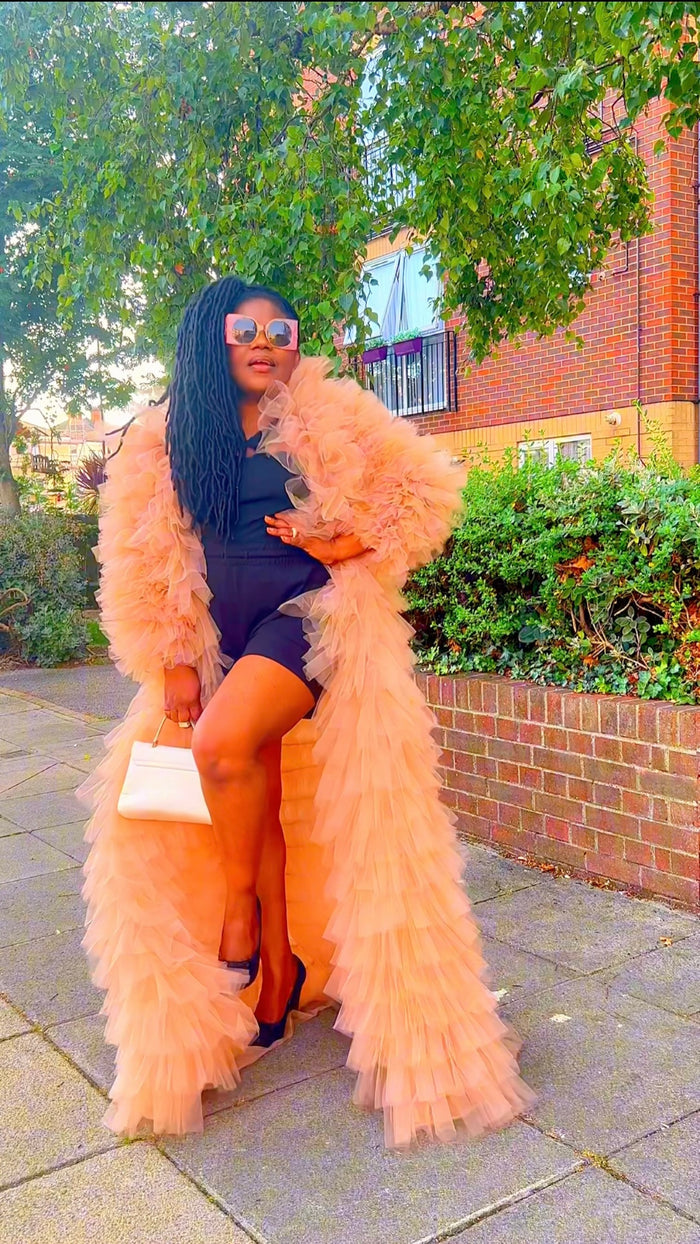 Woman in a long, fluffy peach coat sitting outdoors on a sidewalk.