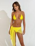A woman stands against a plain white background, showcasing a bright yellow bikini complemented by a sheer TIGERMIST PEACHY SARONG in neon yellow, elegantly tied at her waist. Her hair cascades down as she gazes slightly downward, one arm resting by her side and the other gently touching the sarong.