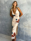 A woman stands against a textured grey wall, wearing the Percy Maxi Dress by Poppy Butterfly. The sleeveless, bodycon fit dress features a deep V-neckline and a side slit. The cream-colored maxi dress showcases a paisley print with blue and rust accents. Her blonde hair is styled in loose waves.