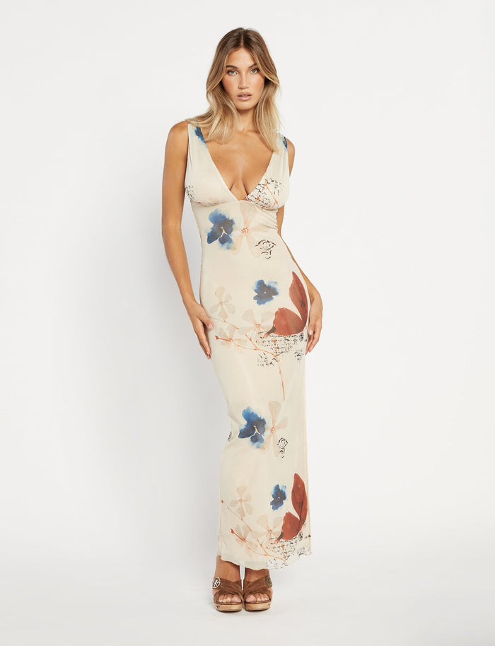 A woman stands against a textured grey wall, wearing the Percy Maxi Dress by Poppy Butterfly. The sleeveless, bodycon fit dress features a deep V-neckline and a side slit. The cream-colored maxi dress showcases a paisley print with blue and rust accents. Her blonde hair is styled in loose waves.