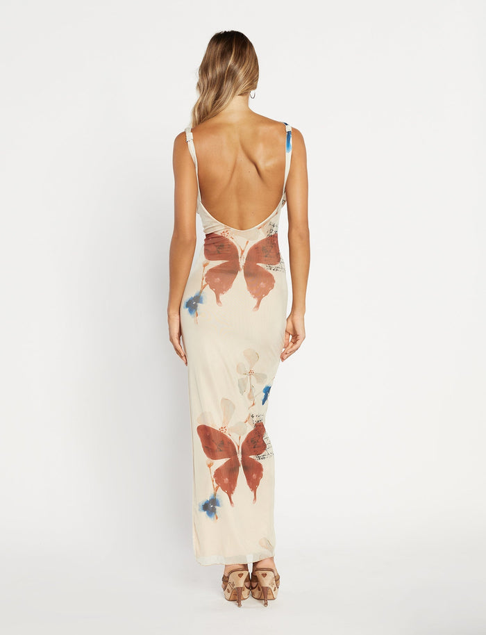 A woman stands against a textured grey wall, wearing the Percy Maxi Dress by Poppy Butterfly. The sleeveless, bodycon fit dress features a deep V-neckline and a side slit. The cream-colored maxi dress showcases a paisley print with blue and rust accents. Her blonde hair is styled in loose waves.
