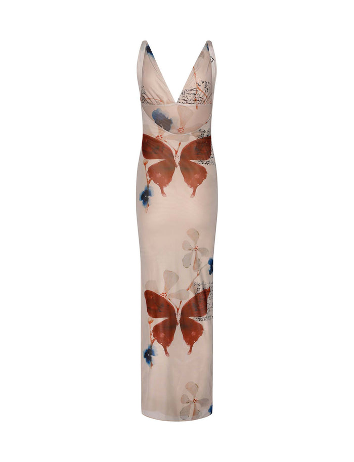 A woman stands against a textured grey wall, wearing the Percy Maxi Dress by Poppy Butterfly. The sleeveless, bodycon fit dress features a deep V-neckline and a side slit. The cream-colored maxi dress showcases a paisley print with blue and rust accents. Her blonde hair is styled in loose waves.