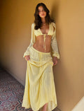 A person with long, wavy hair is standing in a hallway. They are wearing a yellow two-piece outfit from TIGERMIST, featuring the PHILLIPA SKIRT - YELLOW : LEMON and a cropped tie-front top. Their hand rests gently on the skirt, and the floor displays a decorative tile pattern.