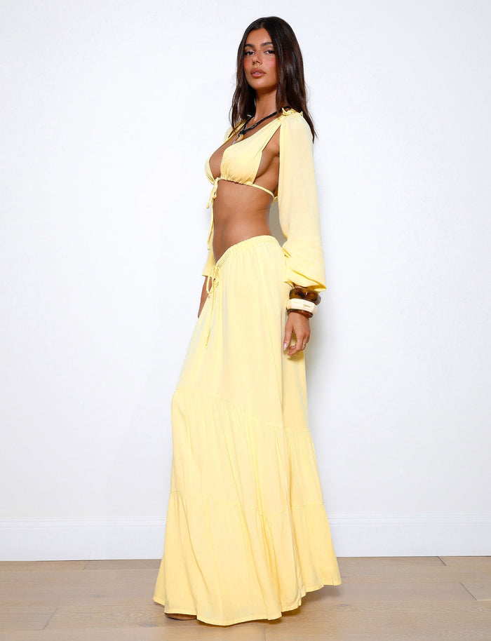 A woman with long dark hair stands against a plain white background, showcasing the PHILLIPA SKIRT - YELLOW : LEMON by TIGERMIST. The light yellow two-piece outfit includes a crop top with full sleeves and a long, flowing skirt. She faces to the side, revealing a portion of her midriff in the stylish attire.