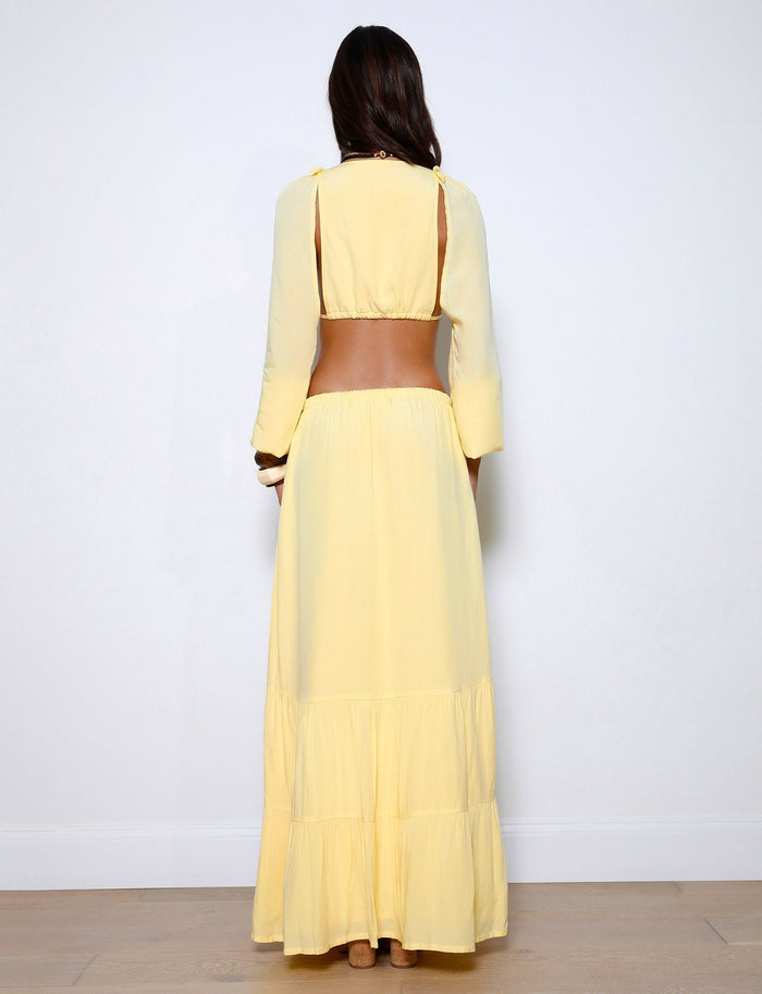A woman with long hair is standing against a white background, facing away from the camera. She is wearing the PHILLIPA SKIRT - YELLOW : LEMON by TIGERMIST, which features tiered layers near the bottom and a brown belt-like detail around her waist.
