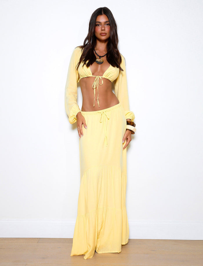 A woman stands against a plain white background wearing a matching yellow outfit from TIGERMIST, featuring the PHILLIPA SKIRT - YELLOW: LEMON and a long-sleeved cropped top. She poses with her hands relaxed by her sides, gazing directly at the camera.