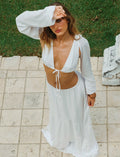 A woman dressed in the TIGERMIST PHILLIPA TOP - WHITE and matching bottom stands on a tiled patio. She is looking up with one hand shielding her eyes from the sun. Her top features long sleeves and ties in the front. The scene is bright with greenery in the background.