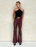 A woman stands against a light beige wall, wearing a black halter top and high-waisted, cherry red leather pants from Tiger Mist. Her hair is styled in a loose updo, with a few strands framing her face. She completes the look with black patent leather pointed-toe shoes.