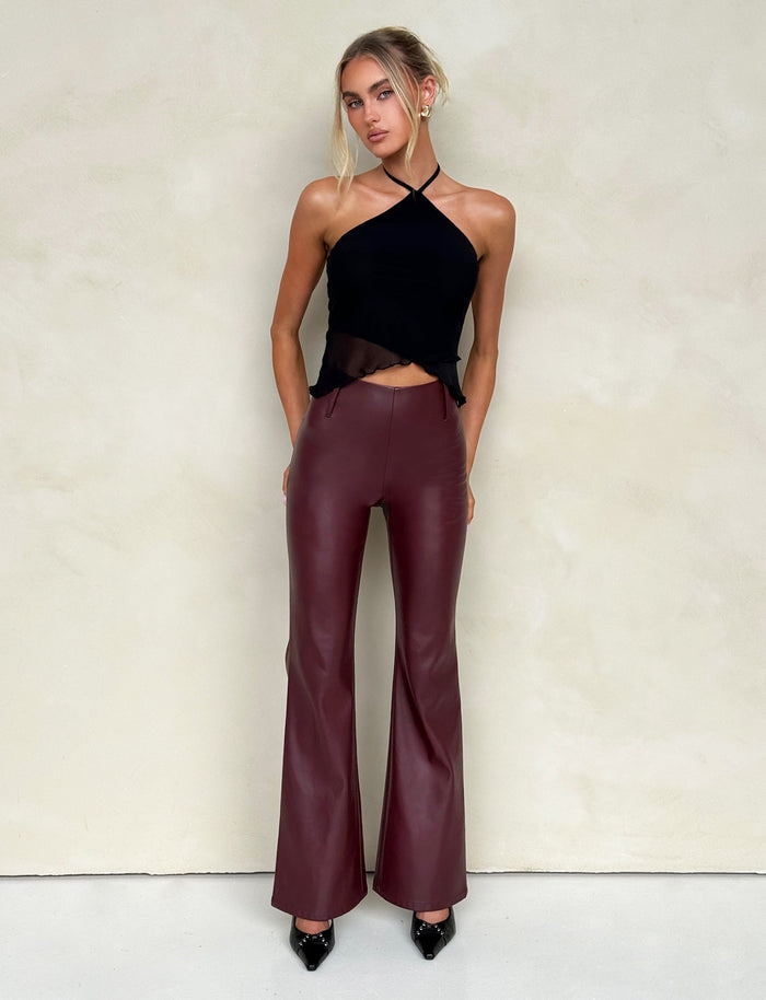 A woman stands against a light beige wall, wearing a black halter top and high-waisted, cherry red leather pants from Tiger Mist. Her hair is styled in a loose updo, with a few strands framing her face. She completes the look with black patent leather pointed-toe shoes.