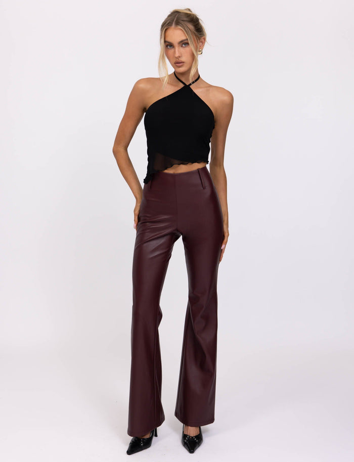 A woman stands against a plain white background, wearing a black halter top and the PU TEDDIE PANT in Cherry Red from TIGERMIST. She has her hair styled in a casual updo and is wearing black pointed-toe heels. She strikes a confident pose with one hand on her hip.