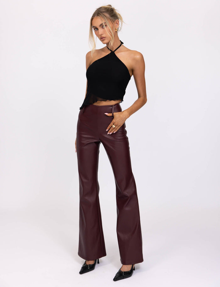 A woman stands against a plain white background wearing a sleeveless, black halter top and high-waisted PU Teddie Pants in cherry red with a slight flare. She has blonde hair tied back and is posing with one hand on her hip and the other hanging by her side. Her striking outfit from TIGERMIST is completed with black heels.