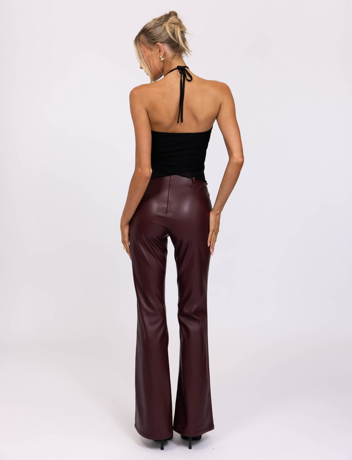 A blonde-haired woman is wearing a sleeveless, black halter top and the high-waisted PU TEDDIE PANT in Cherry Red faux leather with flared legs from TIGERMIST. She stands facing away from the camera against a plain white background.