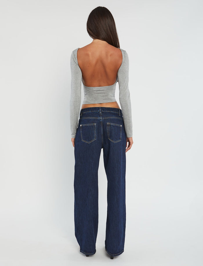 A person with long brown hair is shown from the back, dressed in a chic TIGERMIST outfit. They are wearing the QUINNELL TOP - GREY, a long-sleeved, backless top paired with dark blue wide-leg jeans. The background is plain white.