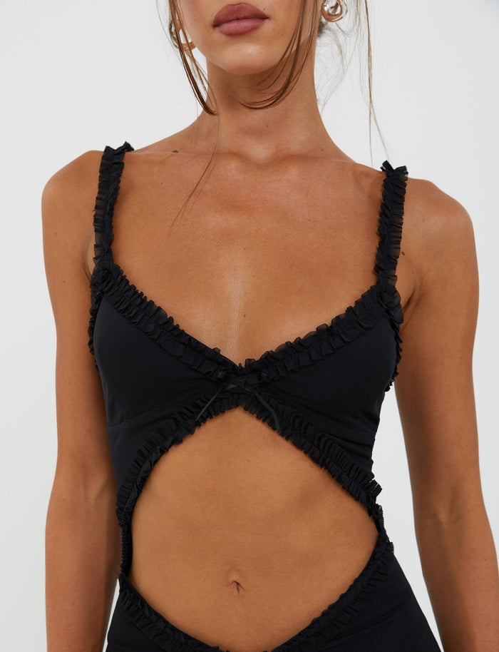 A woman is wearing the Rayla Dress in black from TigerMist, featuring a deep V neckline and cut-out midsection with crisscross detailing on the chest. Her hair is tied back, with tendrils framing her face. Only her upper body and part of her face are visible against a plain background.