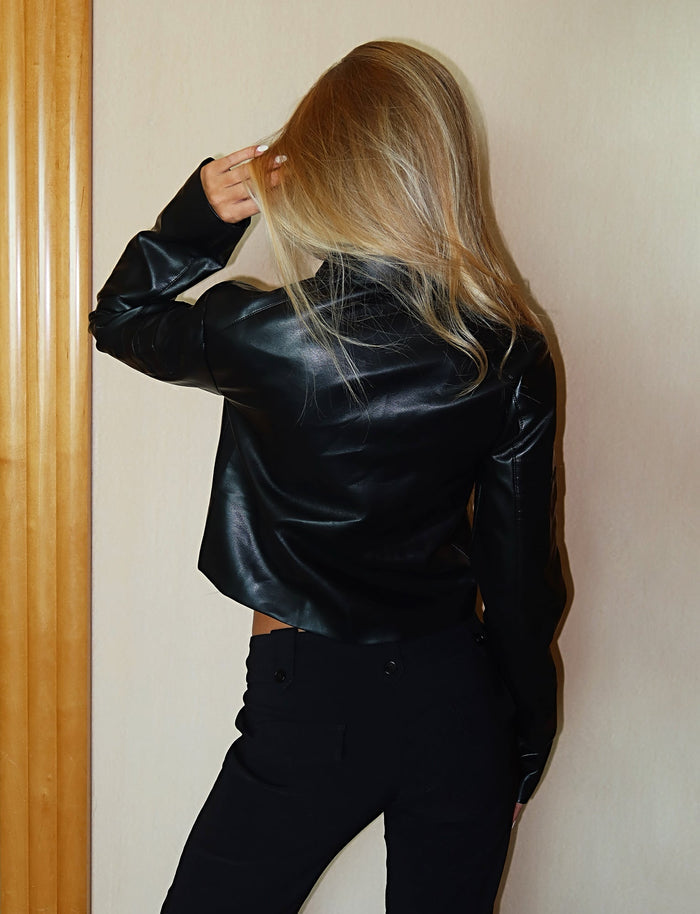 A person with long blonde hair stands with their back to the camera, wearing TIGERMIST's RYDER JACKET - BLACK and black pants. One hand is raised to their head, lightly touching their hair. The background features a light-colored wall with wooden trim on the left.