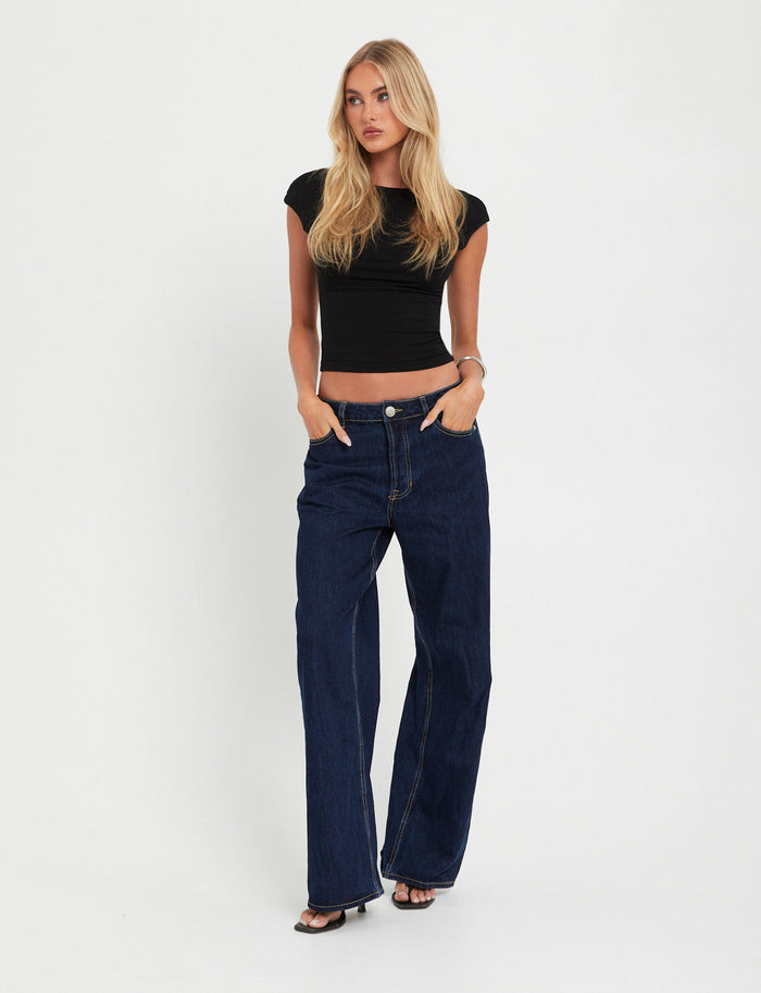 A person with long blonde hair stands against a plain white background, wearing the fitted RYLAN TOP - BLACK and high-waisted dark blue wide-leg jeans from TigerMist. They have their hands in their pockets and are wearing open-toed black shoes.
