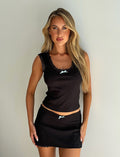 A woman with long blond hair stands against a plain wall, wearing the SABINE SKORT in black from TIGERMIST, adorned with delicate lace trim and small blue bows. She looks directly at the camera with a neutral expression, her hands resting on her hips.