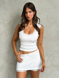 A woman with long, wavy brown hair stands against a textured gray wall. She is wearing the SABINE TOP - WHITE from TIGERMIST, a sleeveless fitted top with lace trim, along with a matching short skirt. With a neutral expression, her left hand rests on her hip.