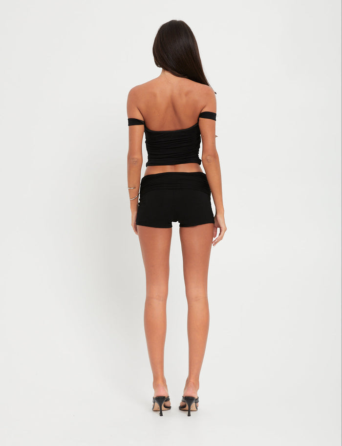 A person with long dark hair, facing away from the camera, is dressed head to toe in TIGERMIST. They are wearing the black SAFAA TOP along with matching black shorts and high heels against a plain white background.