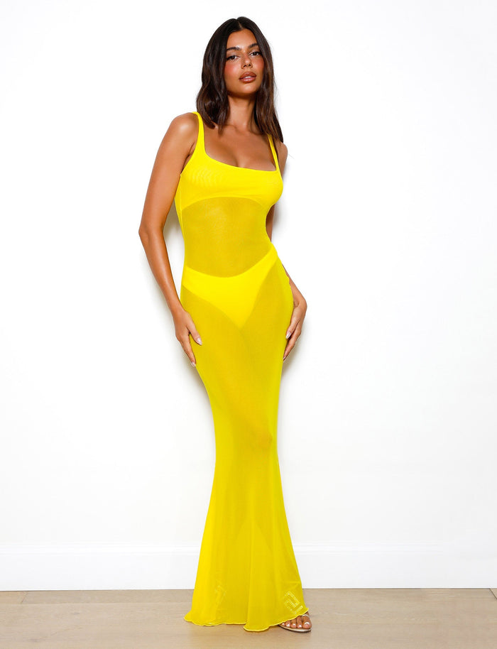 A woman with long dark hair stands against a plain white background, wearing the SAMIR DRESS - YELLOW from TIGERMIST. The fitted, semi-transparent dress in bright neon yellow extends to the floor. She poses with one hand on her hip, looking directly at the camera while wearing open-toed shoes.