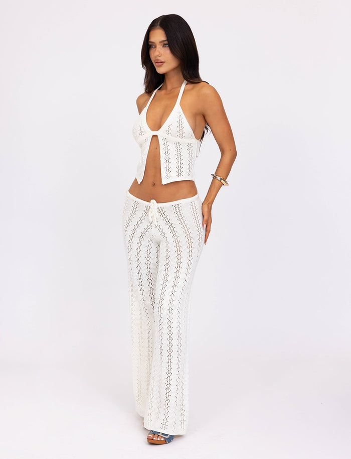 A woman with long, dark hair is wearing the white crochet halter top and matching SANDOR PANT - WHITE from TIGERMIST. She stands against a plain white background, posing with one hand on her hip and the other at her side. She accessorizes with a bracelet and blue sandals.