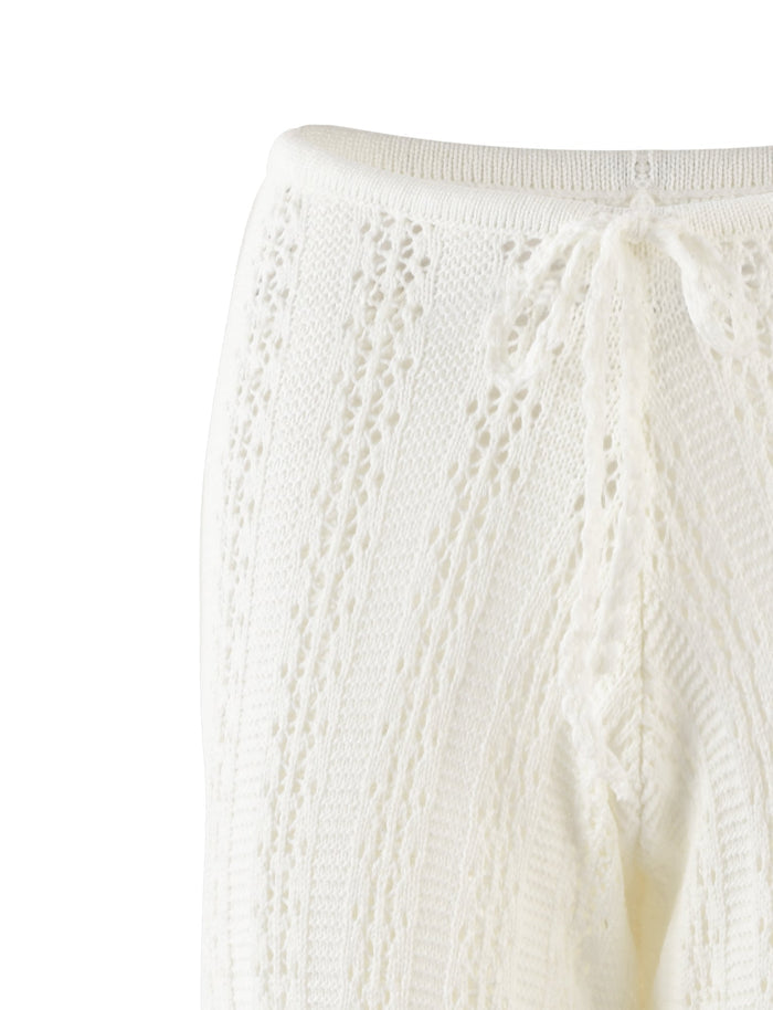 Close-up image of the SANDOR PANT in white from TIGERMIST. The fabric showcases a detailed textured lace design with vertical patterns and a decorative bow tied near the waistband, giving the garment an elegant and delicate appearance.