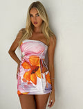 A woman with long blonde hair wearing the Sariah Top - Multi with a Macro Floral design by TIGERMIST stands against a white textured wall. She gazes directly at the camera, her arms relaxed by her sides. The top features vibrant colors, including orange and pink flowers.