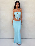 A woman with long dark hair stands against a plain background, wearing a pastel blue, two-piece knit outfit from TIGERMIST. The SHAH SKIRT is paired with a matching top that features a bow-like design with a halter neck. She completes her look with high-heeled sandals and wears a bracelet on her right wrist.