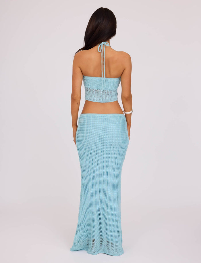 A woman is seen from the back wearing a light blue, knit halter top and the SHAH SKIRT in pale blue from TIGERMIST. The top ties at the neck and exposes her midriff, while the skirt is long and flowy. She has long, dark hair and stands against a plain, light-colored background.