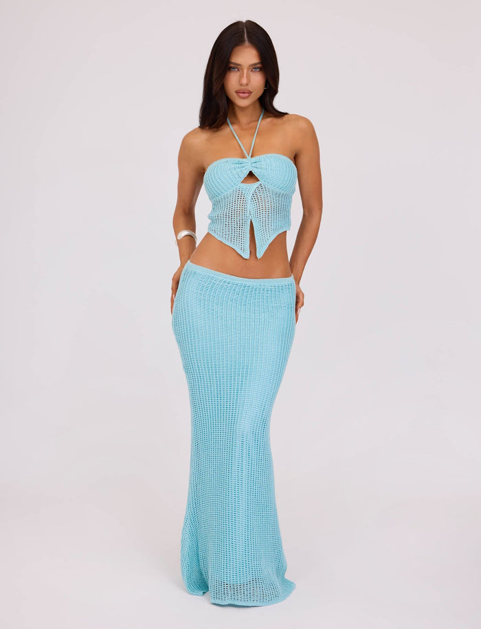 A woman stands facing the camera, displaying the TIGERMIST SHAH TOP in a pastel blue crochet style with a knot detail at the bust, paired with a matching long skirt. She has long dark hair and a neutral expression, posing against a plain white background.