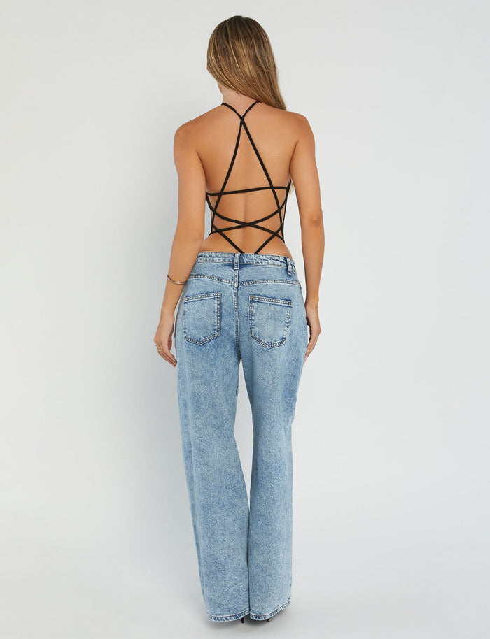 A woman stands with her back to the camera, showcasing a chic Tiger Mist Shauna Bodysuit in black and high-waisted, light blue denim jeans. Her long, light brown hair cascades down against a plain, light-colored background.