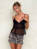 A woman is posing against a plain backdrop in a stunning Tiger Mist outfit. She wears the SINGE TOP - BLACK from TIGERMIST, featuring a black lace camisole with thin straps and a sheer midsection, perfectly paired with leopard-print shorts. Her hair is styled in a loose updo with strands framing her face, and she has stud earrings in her ears.
