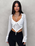 A woman with long dark hair is wearing the SOPHA TWO PIECE TOP - WHITE from TIGERMIST paired with black pants. She accessorizes with a silver heart-shaped necklace and has a confident expression, standing against a neutral gray background.
