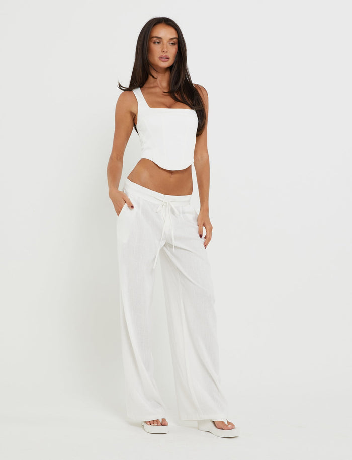 A woman stands against a plain white background, styled in a chic TIGERMIST SULLIVAN TOP - WHITE and loose trousers with pockets. With long dark hair and hands casually tucked in her pockets, she looks directly at the camera. She completes her look with white sandals.