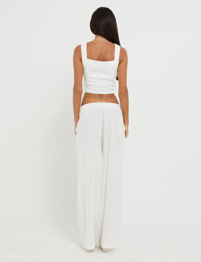 A person with long dark hair is standing with their back to the camera, showcasing a stylish outfit from TIGERMIST. They are wearing the SULLIVAN TOP - WHITE, a white sleeveless crop top, paired with loose-fitting white pants, all set against a plain white background.