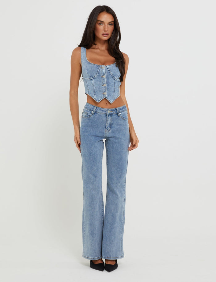 A person with long dark hair is standing against a plain white background, confidently wearing a light blue denim corset top and the TATYANA JEANS - BLUE : LIGHT WASH : LIGHT DENIM from TIGERMIST, paired with black shoes. The individual has a poised stance with hands by their sides.