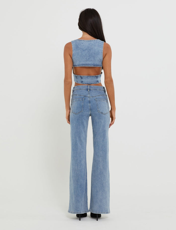 A woman with long dark hair stands facing away from the camera. She wears a stylish sleeveless denim crop top with a cut-out back and high-waisted, flared TATYANA JEANS in light blue wash from TIGERMIST. The background is plain white, and she is standing on black heeled shoes.