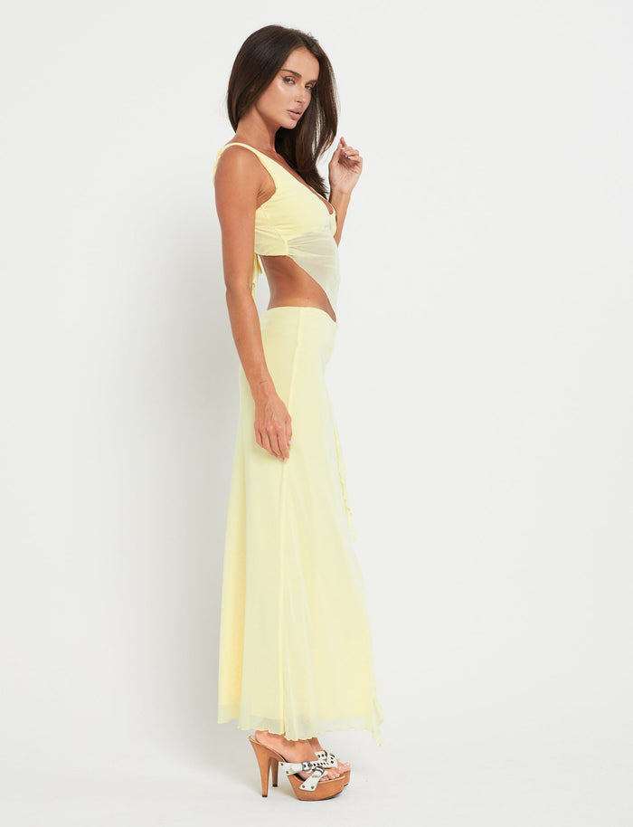 A woman with long dark hair poses against a plain white background, wearing the TIGERMIST THEO SKIRT - YELLOW : LEMON, a light yellow, sleeveless cut-out dress with a flowing skirt. She pairs it with wooden platform heels featuring a white and black spotted design. She gazes to her right while gently holding her hair with her left hand.