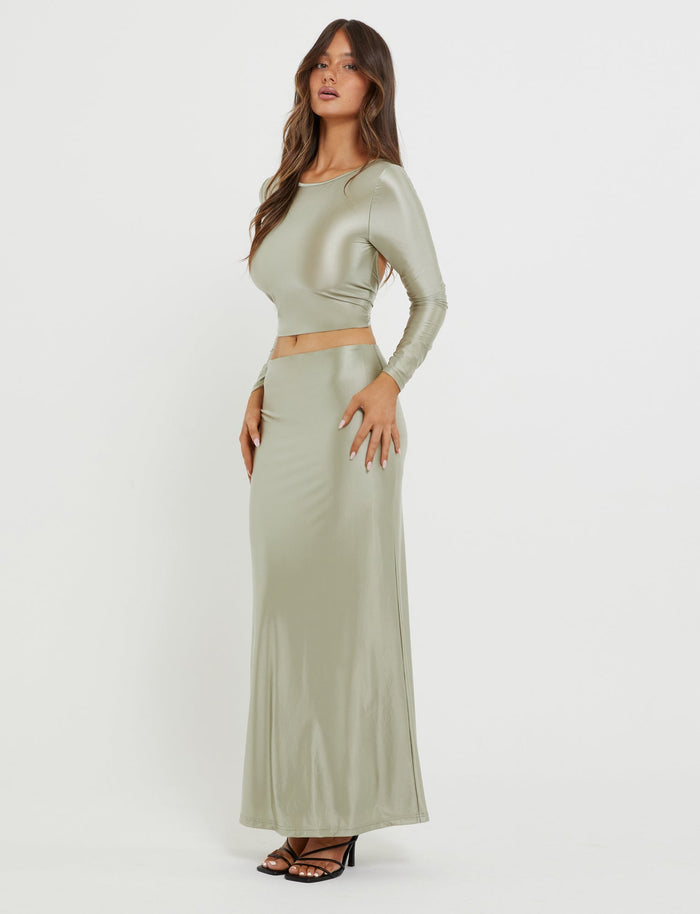 A woman with long brown hair is wearing a shiny, form-fitting, Matcha Green Thessaly Top from Tiger Mist. She pairs it with a coordinating high-waisted, floor-length skirt and black high-heeled sandals. She stands against a plain white background with one hand on her hip.